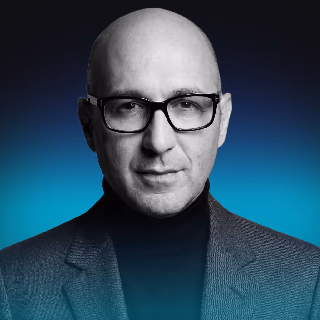 Portrait of a bald man wearing black glasses, a dark turtleneck, and a gray blazer against a blue gradient background.
